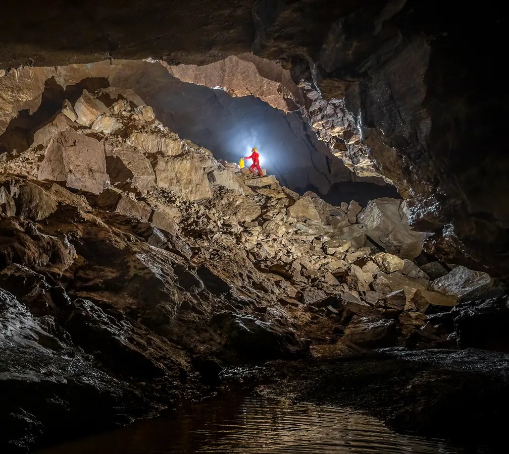Spéléologue en combinaison rouge au centre d’un vaste éboulis, dans une grande salle souterraine.