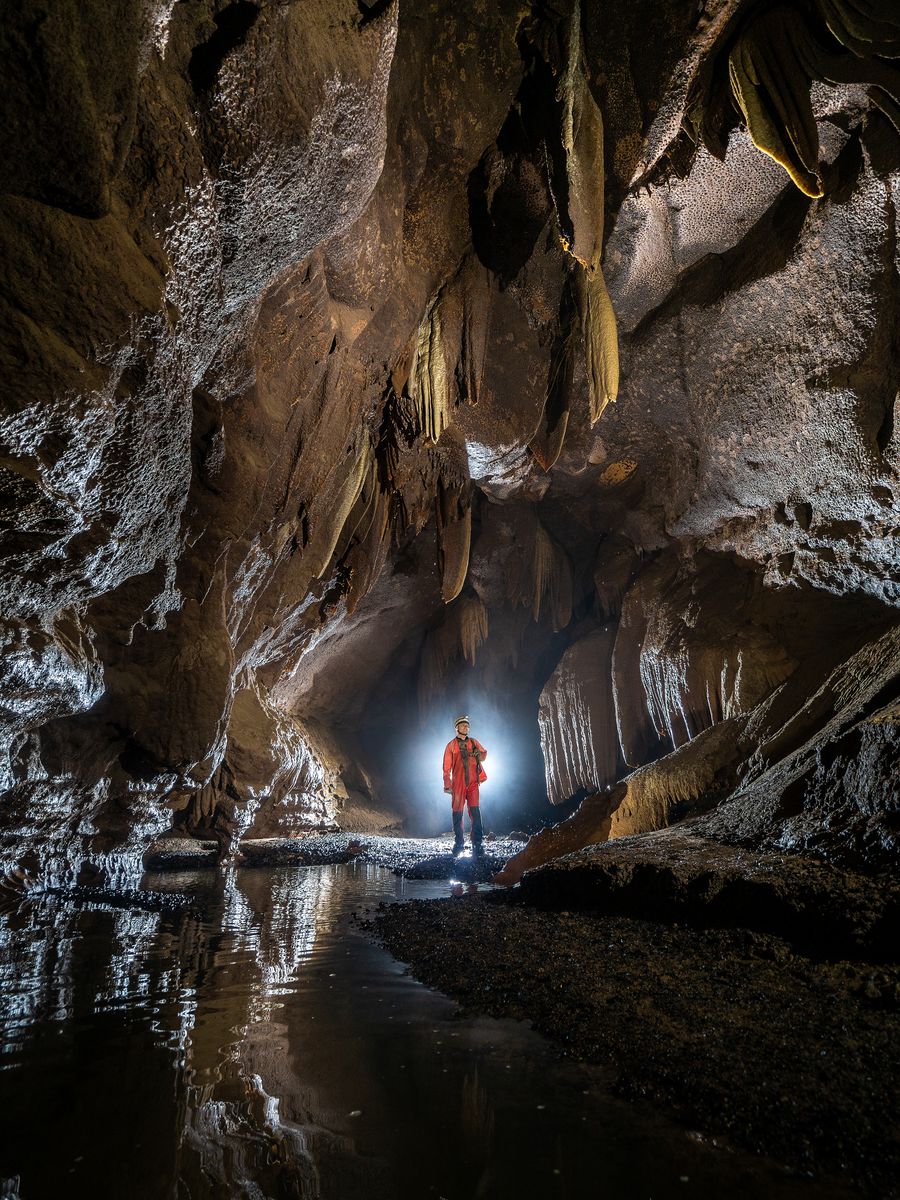 Spéléologue debout au bord d'un ruisseau souterrain, sous des draperies et des stalactites.