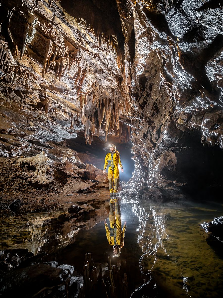 Spéléologue en combinaison jaune se reflétant dans un plan d'eau, sous un plafond couvert de concrétions.