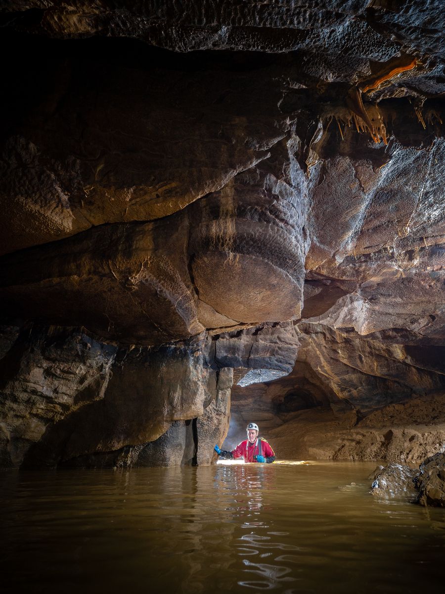 Spéléologue en combinaison étanche avançant dans une rivière souterraine, de l'eau jusqu'à la taille.