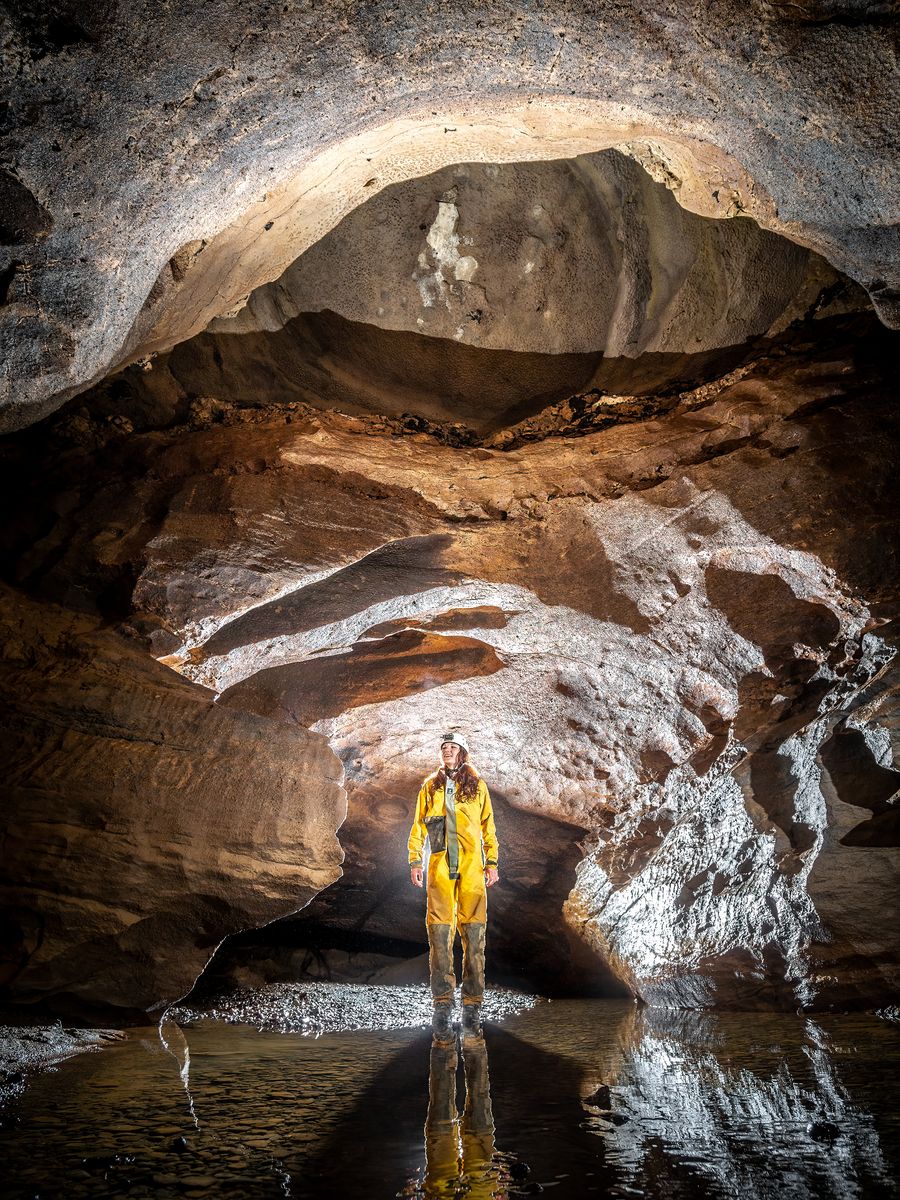 Spéléologue en combinaison jaune sous une voûte de roche stratifiée, au bord d'une vasque d'eau.